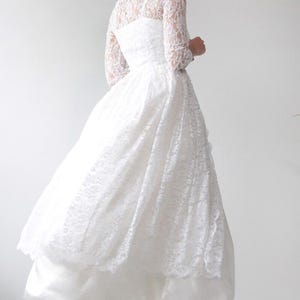 May include: A vintage white wedding dress with a lace bodice and long sleeves. The full skirt is layered, and the dress is paired with light blue sneakers. The dress is a classic bridal style.