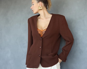 May include: A brown blazer with a V-neck and three buttons. The blazer has a tailored fit and features two front pockets. The model is wearing a patterned top underneath and gold earrings. The blazer is a classic piece of clothing.
