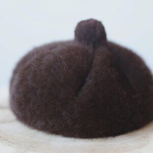 May include: A dark brown beret with a small pom-pom on top. The hat is made of a soft, fuzzy material and sits on a light-colored surface. This classic beret is a stylish accessory.