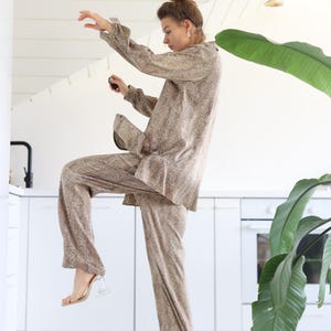 May include: A neutral-toned two-piece pajama set with a textured pattern. The long-sleeved top and wide-legged trousers are paired with clear heeled sandals. The setting is a modern kitchen with white cabinets and a large green plant.