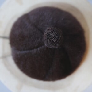 May include: A dark brown beret with a small pom-pom on top, set inside a light-colored, textured bowl. The beret has a soft, fuzzy texture and a rounded shape, suggesting a warm, winter accessory.