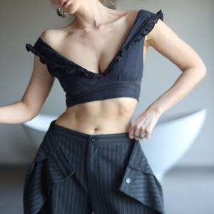 Op de afbeelding: Een zwarte off-the-shoulder crop top met ruches en een broek met hoge taille en krijtstreep. De broek heeft unieke zakdetails. De outfit is een moderne interpretatie van klassieke kleermakerij.