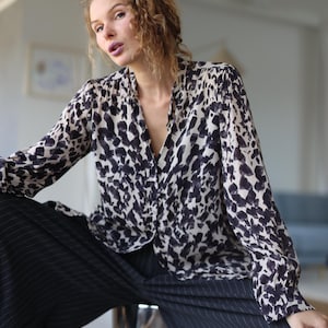 Op de afbeelding: Een blouse met lange mouwen en knoopsluiting op een crèmekleurige basis met een abstract donkerblauw penseelstreekpatroon, gecombineerd met een donker antracietgrijze pantalon met krijtstreep en wijde pijpen. Zachte kraag en geplooide schouders.