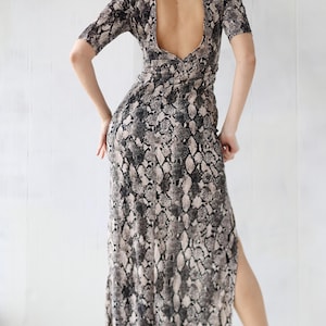 Könnte beinhalten: Maxikleid mit Schlangenmuster in Grau-, Schwarz- und Beigetönen. Das Kleid hat kurze Ärmel, einen Schlüsselloch-Rücken und einen hohen Schlitz. Das Model trägt transparente Absätze.
