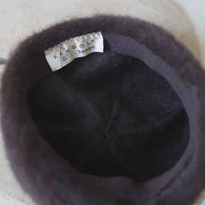 May include: A dark brown Kangol design hat, viewed from the inside. The hat has a soft, fuzzy texture and a dark brown interior lining. A white tag with the Kangol logo and "Made in England" is visible.