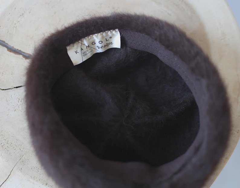 May include: A dark brown Kangol design hat, viewed from the inside. The hat has a soft, fuzzy texture and a dark brown interior lining. A white tag with the Kangol logo and "Made in England" is visible.