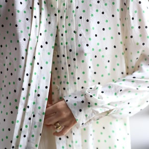 Op de afbeelding: Een overhemd met lange mouwen in een off-white kleur met een herhalend patroon van kleine zwarte en felgroene stippen. Het overhemd heeft een ronde halslijn met verticale plooien langs de knoopsluiting en manchetten met knopen.