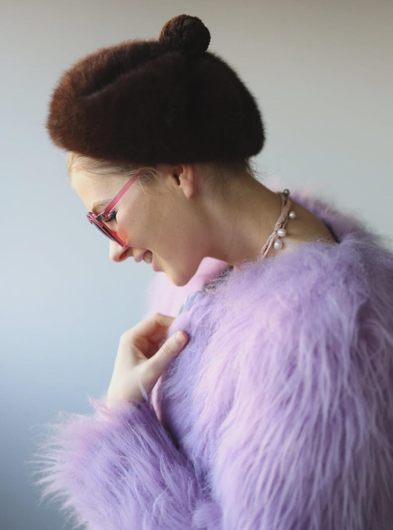 May include: A woman wearing a brown fur beret with a small ball on top, pink-framed sunglasses, a pearl necklace, and a fluffy lavender coat. The coat has long, soft fur. The woman is looking down and smiling.