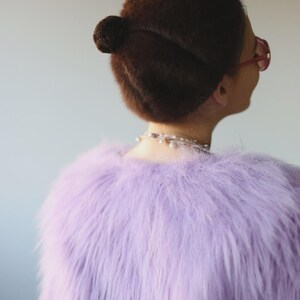 May include: A person's back view, wearing a light purple faux fur jacket. The person has dark brown hair styled in a bun and is wearing pink-framed glasses and a pearl necklace. The background is a solid light gray.