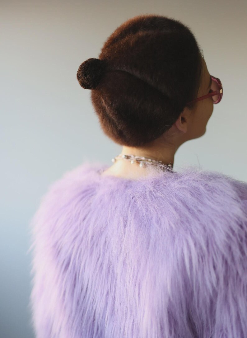 May include: A person's back view, wearing a light purple faux fur jacket. The person has dark brown hair styled in a bun and is wearing pink-framed glasses and a pearl necklace. The background is a solid light gray.