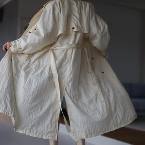May include: A cream-coloured trench coat featuring a textured fabric, a belted waist, a back storm flap, and dark brown buttons. The coat has long sleeves with adjustable cuffs and a flared hemline that falls below the knees.