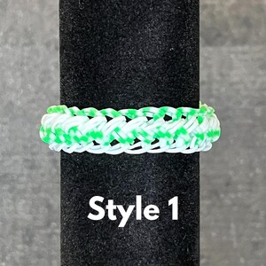 May include: A white and green rubber band bracelet with a woven pattern. The bracelet is on a black display stand. The text "Style 1" is printed below the bracelet.
