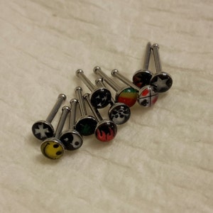 May include: A collection of nose studs with various designs, including a smiley face, yin-yang symbol, and a star. The studs have a silver-colored post and a round, decorative top with colorful graphics.