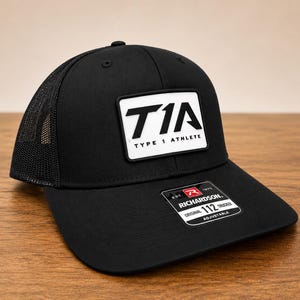 May include: A black trucker hat with a mesh back and a rectangular white patch on the front featuring the black text T1A TYPE 1 ATHLETE. A black and white sticker on the brim displays the text RICHARDSON ORIGINAL 112 TRUCKER EST 1970 ADJUSTABLE.