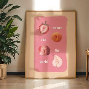 Puede incluir: Una impresión artística enmarcada con un fondo rosa y el texto "women run the world". La impresión incluye ilustraciones de una fresa cortada, un melocotón, un higo y una pera. La obra de arte se exhibe en una habitación con una gran planta en maceta.