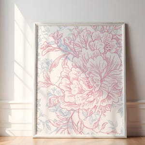 May include: A framed floral print featuring a large pink peony design with blue and green accents on a cream background. The artwork is displayed against a white wall and wooden floor, with natural light illuminating the scene.