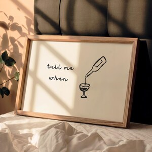 Puede incluir: Un cuadro enmarcado con un dise&ntilde;o minimalista. La obra de arte presenta una botella de vino vertiendo en una copa, con las palabras "tell me when" en escritura negra. El marco es de madera clara.