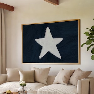 May include: A living room with a large framed artwork featuring a white star on a dark blue background. A beige sofa with several pillows sits in front of the artwork. A glass coffee table and a wooden chair are in the foreground. The room has light-colored walls, a window with sheer curtains, and a plant in the corner.