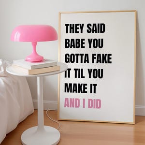 Può includere: Una lampada rosa brillante a forma di fungo è appoggiata su un tavolino bianco con dei libri. Una stampa incorniciata con il testo "THEY SAID BABE YOU GOTTA FAKE IT TIL YOU MAKE IT AND I DID" è appoggiata al muro.