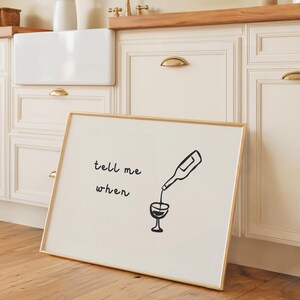 May include: A framed art print with a gold frame leans against a kitchen cabinet. The print features a black illustration of a wine bottle pouring into a wine glass, with the words 'tell me when' above the illustration. The kitchen has white cabinets with gold handles and a wooden countertop.