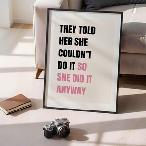 May include: A framed print with the text "THEY TOLD HER SHE COULDN'T DO IT SO SHE DID IT ANYWAY" in black and pink. A vintage camera and a brown book are in front of the print. The print is leaning against a wall.