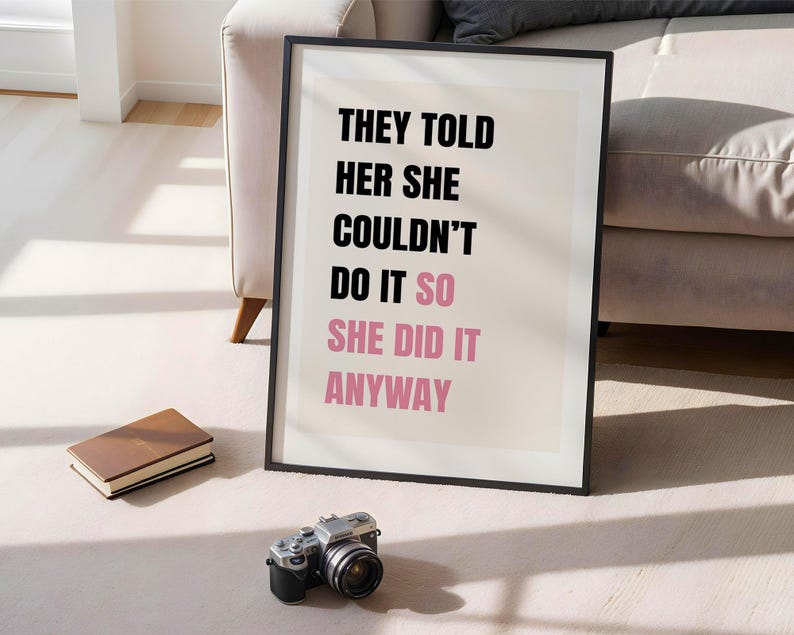 May include: A framed print with the text "THEY TOLD HER SHE COULDN'T DO IT SO SHE DID IT ANYWAY" in black and pink. A vintage camera and a brown book are in front of the print. The print is leaning against a wall.