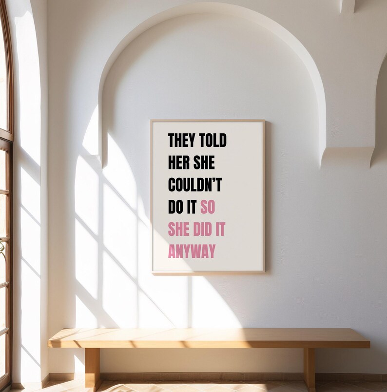 May include: A framed print with the text "THEY TOLD HER SHE COULDN'T DO IT SO SHE DID IT ANYWAY" in black and pink. The print hangs on a white wall above a wooden bench. The room has an arched doorway.