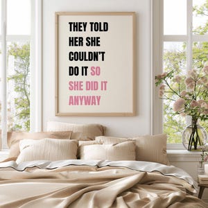 May include: A framed print with the text "THEY TOLD HER SHE COULDN'T DO IT SO SHE DID IT ANYWAY" in black and pink. The print hangs above a bed with beige bedding and pillows. A vase of flowers sits on a small table.