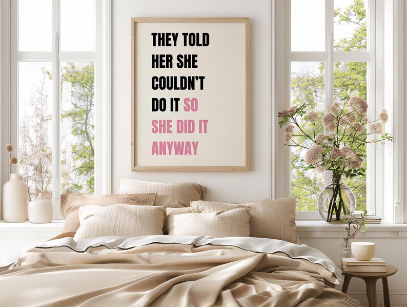 May include: A framed print with the text "THEY TOLD HER SHE COULDN'T DO IT SO SHE DID IT ANYWAY" in black and pink. The print hangs above a bed with beige bedding and pillows. A vase of flowers sits on a small table.