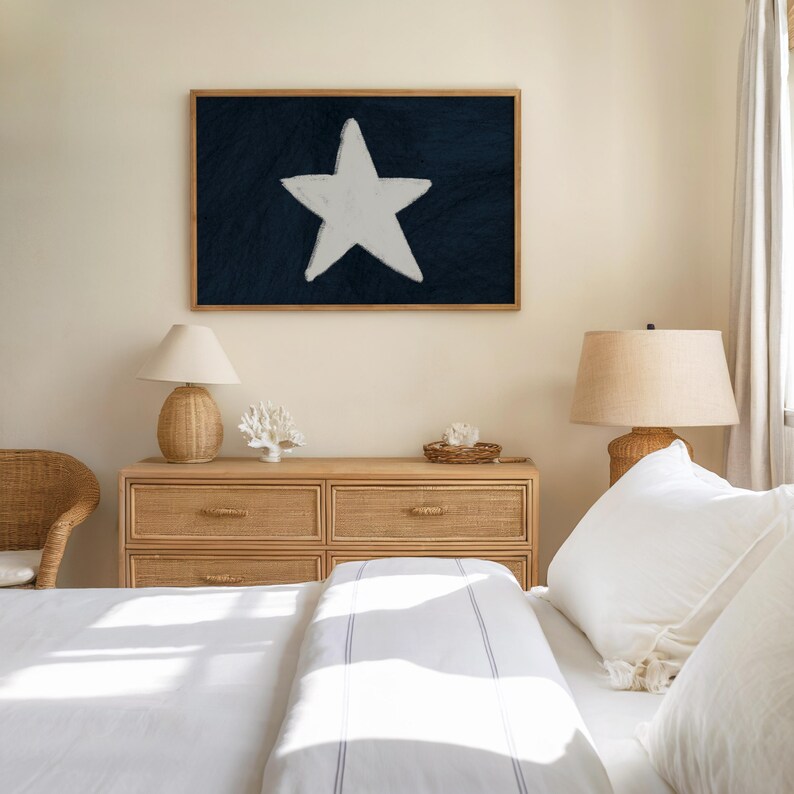 May include: A framed artwork featuring a large white star on a dark blue background. The artwork is displayed above a light wood dresser with a lamp, coral decor, and a woven basket. The room has a neutral color palette with white bedding.