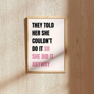 May include: A framed print with the text "THEY TOLD HER SHE COULDN'T DO IT SO SHE DID IT ANYWAY." The text is in black and pink on a white background, set in a light wood frame. The print is mounted on a light wood-grain wall.
