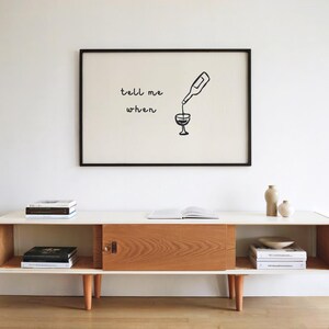 Puede incluir: Una impresi&oacute;n art&iacute;stica enmarcada con las palabras "tell me when" y una botella de vino vertiendo en una copa. La impresi&oacute;n cuelga sobre un mueble de estilo moderno de mediados de siglo con libros y objetos decorativos.