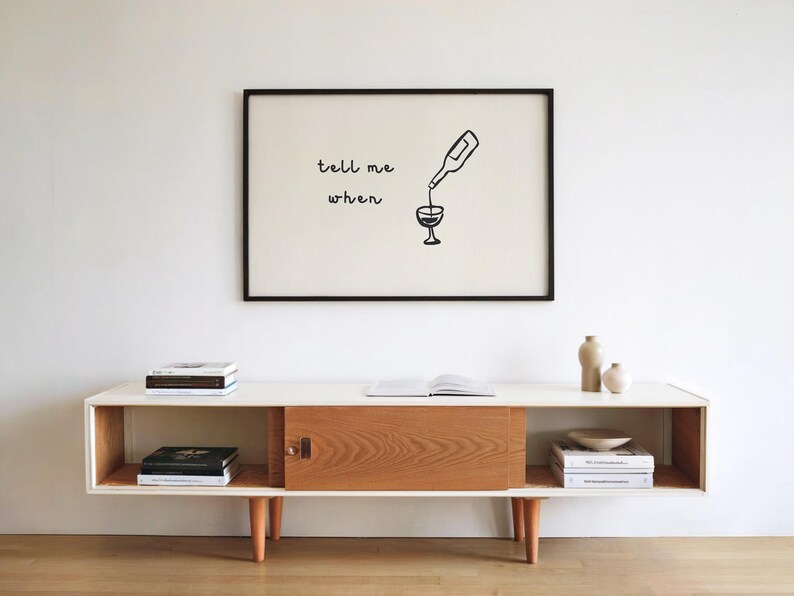 Puede incluir: Una impresi&oacute;n art&iacute;stica enmarcada con las palabras "tell me when" y una botella de vino vertiendo en una copa. La impresi&oacute;n cuelga sobre un mueble de estilo moderno de mediados de siglo con libros y objetos decorativos.