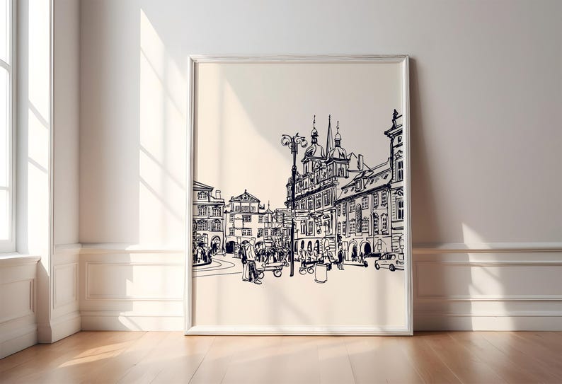May include: A framed black and white sketch of a European city square, featuring detailed architectural drawings of buildings, people, and vehicles. The artwork is displayed on a light beige background, with a white frame.