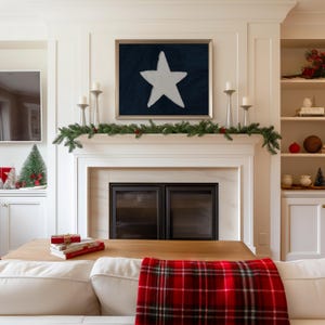 May include: A framed artwork featuring a white star on a dark blue background hangs above a fireplace. The mantel is decorated with greenery, candles, and a red berry garland. A red and black plaid blanket is draped over a white sofa.