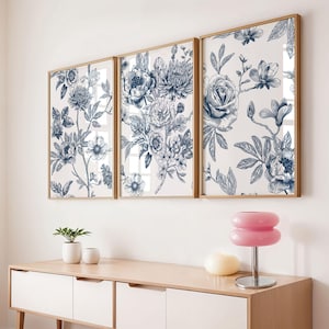 Pu&ograve; includere: Tre stampe artistiche floreali incorniciate con un design botanico nei toni del blu e del bianco. Le stampe sono esposte sopra un mobile in legno chiaro con una lampada rosa e piccole piante in vaso.