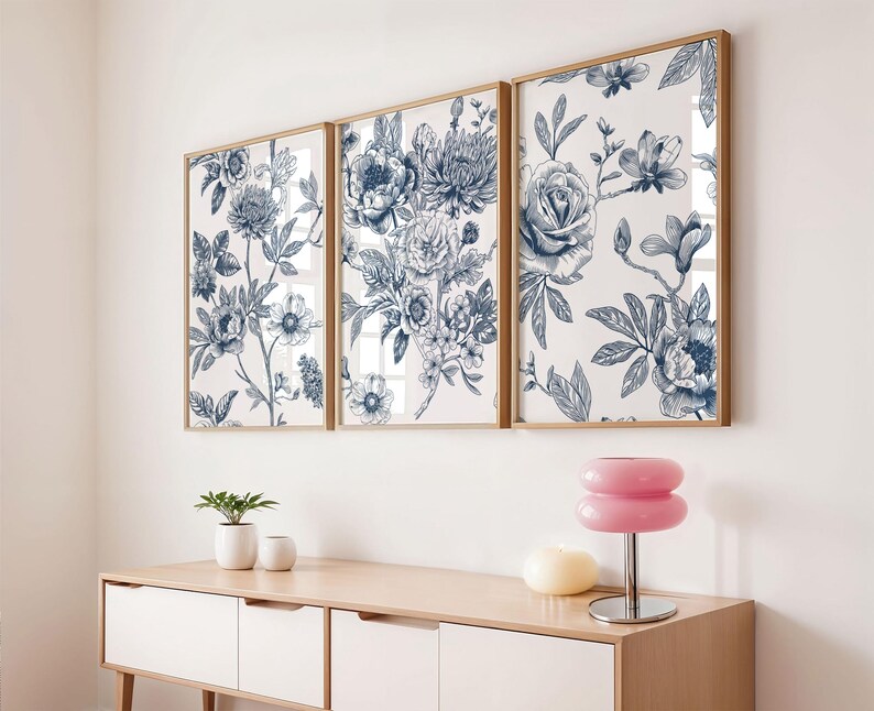 Pu&ograve; includere: Tre stampe artistiche floreali incorniciate con un design botanico nei toni del blu e del bianco. Le stampe sono esposte sopra un mobile in legno chiaro con una lampada rosa e piccole piante in vaso.