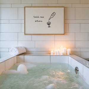 Puede incluir: Una relajante escena de spa con una ba&ntilde;era de azulejos blancos llena de agua y burbujas. Una obra de arte enmarcada con el texto "tell me when" cuelga sobre la ba&ntilde;era. Varias velas blancas est&aacute;n encendidas y una toalla blanca enrollada descansa en el borde.
