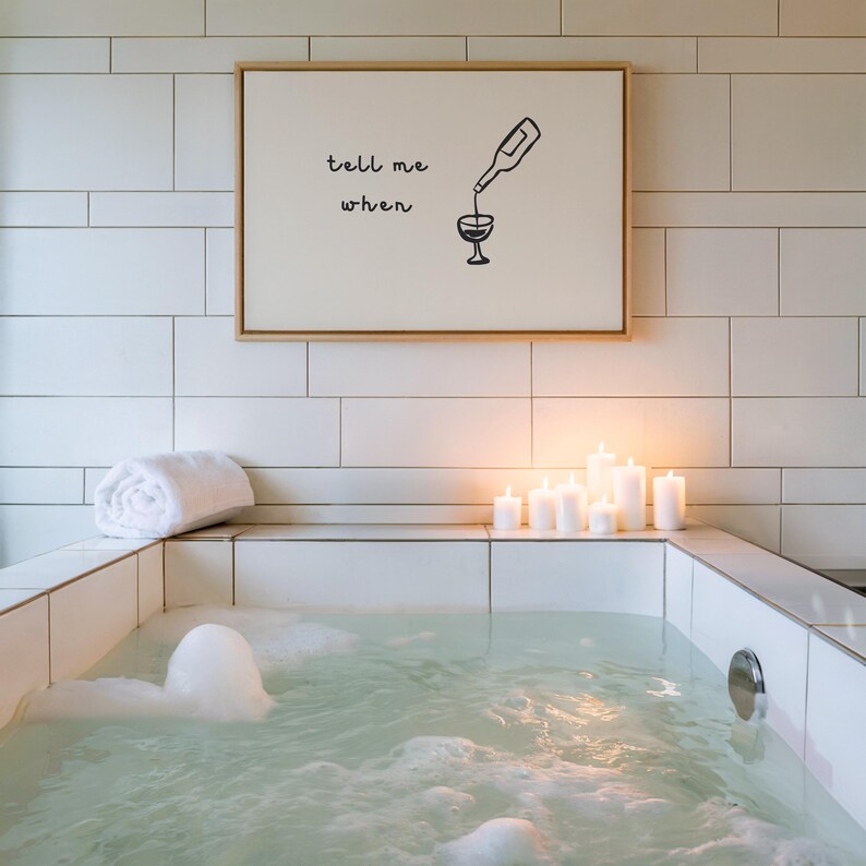 Puede incluir: Una relajante escena de spa con una ba&ntilde;era de azulejos blancos llena de agua y burbujas. Una obra de arte enmarcada con el texto "tell me when" cuelga sobre la ba&ntilde;era. Varias velas blancas est&aacute;n encendidas y una toalla blanca enrollada descansa en el borde.