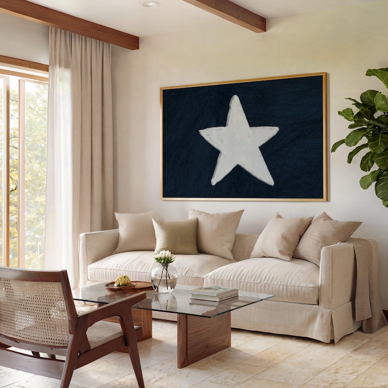May include: A living room with a large framed artwork featuring a white star on a dark blue background. A beige sofa with several pillows sits in front of the artwork. A glass coffee table and a wooden chair are in the foreground. The room has light-colored walls, a window with sheer curtains, and a plant in the corner.