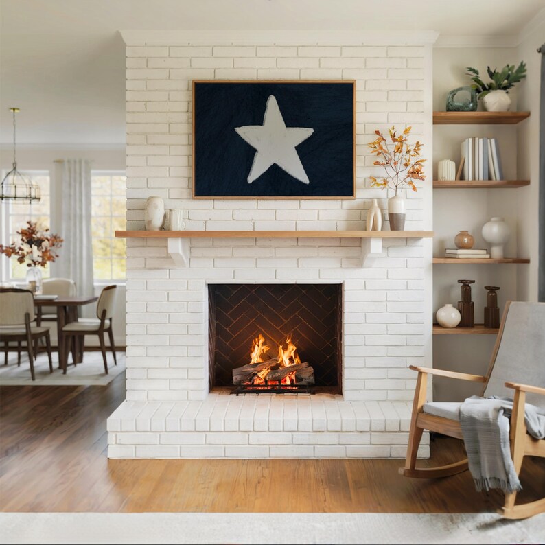 May include: A living room with a white brick fireplace, a wooden mantel, and a framed artwork featuring a white star on a dark blue background. The fireplace has a burning fire inside. Wooden shelves hold decorative items.