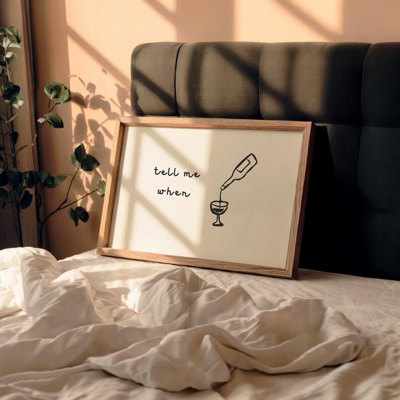 Puede incluir: Un cuadro enmarcado con un dise&ntilde;o minimalista. La obra de arte presenta una botella de vino vertiendo en una copa, con las palabras "tell me when" en escritura negra. El marco es de madera clara.