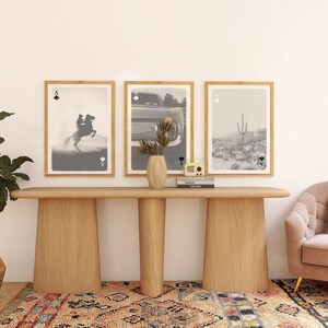 Op de afbeelding: Een moderne woonkamer met drie ingelijste kunstprints met speelkaartenmotieven. Een houten console bevat een vaas, boeken en een kleine radio. Een potplant en een roze bank maken de inrichting compleet.