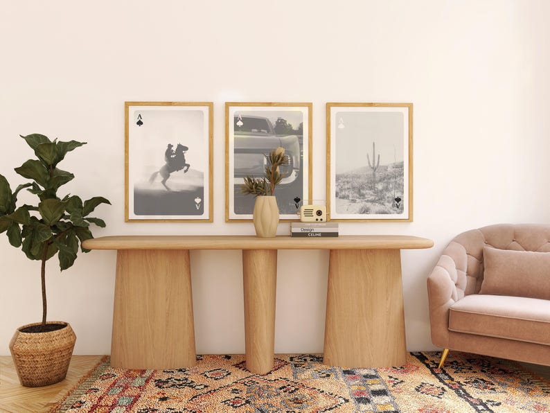 Op de afbeelding: Een moderne woonkamer met drie ingelijste kunstprints met speelkaartenmotieven. Een houten console bevat een vaas, boeken en een kleine radio. Een potplant en een roze bank maken de inrichting compleet.