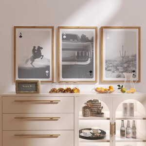 Op de afbeelding: Drie ingelijste zwart-wit prints met houten frames. De prints tonen een cowboy op een paard, een auto en een woestijnlandschap. De Aas van Schoppen staat boven en onder elke print. Een houten bord leest "Welcome".