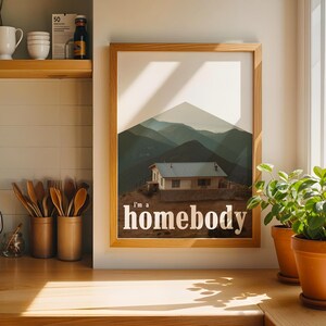 May include: A framed art print featuring a mountain landscape with a small house and the text "I'm a homebody." The print is in a wooden frame and hangs on a white wall. Two potted plants sit on a wooden surface.