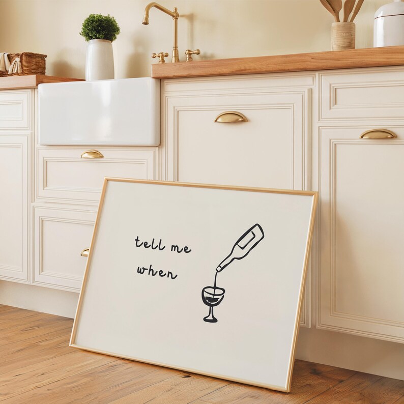 May include: A framed art print with a gold frame leans against a kitchen cabinet. The print features a black illustration of a wine bottle pouring into a wine glass, with the words 'tell me when' above the illustration. The kitchen has white cabinets with gold handles and a wooden countertop.