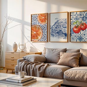 May include: Three framed wall art pieces. One features orange slices, another a blue and white illustration of a coastal town, and the third, red tomatoes. The art is displayed above a gray sofa with decorative pillows.