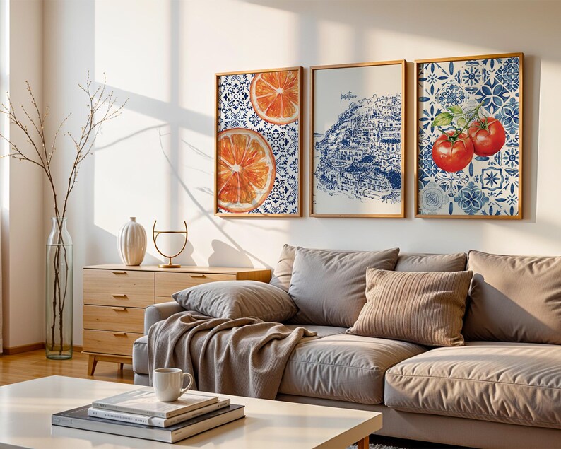 May include: Three framed wall art pieces. One features orange slices, another a blue and white illustration of a coastal town, and the third, red tomatoes. The art is displayed above a gray sofa with decorative pillows.