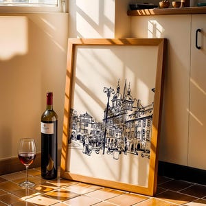 May include: A framed print of a European city scene in black and white, leaning against a wall. A bottle of red wine and a wine glass with red wine are in front of the frame. The frame is made of light brown wood.
